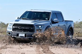 Silver Ford Raptor truck driving through muddy terrain, splashing water.