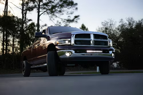 A dark-colored Dodge Ram pickup truck parked on an asphalt road at dusk with its headlights and light bar illuminated.