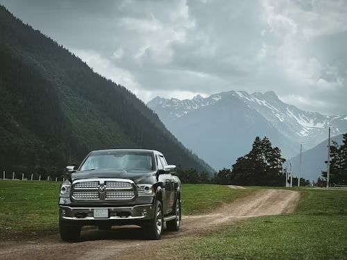 Black truck parked on a dirt road in front of green grass and a mountain range under a cloudy sky.