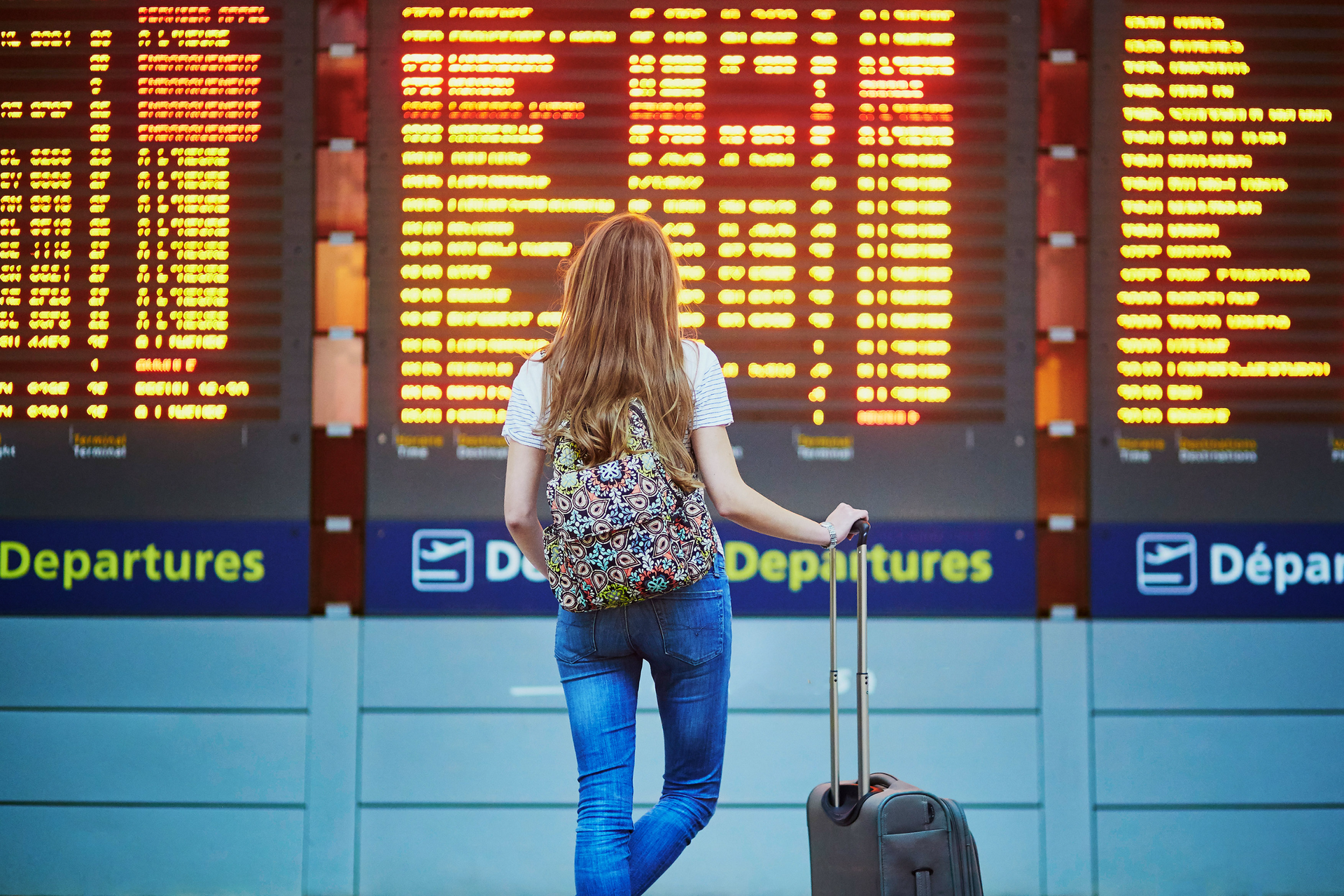 Woman at airport, looking at departure board, pulling a suitcase, waiting to fly.