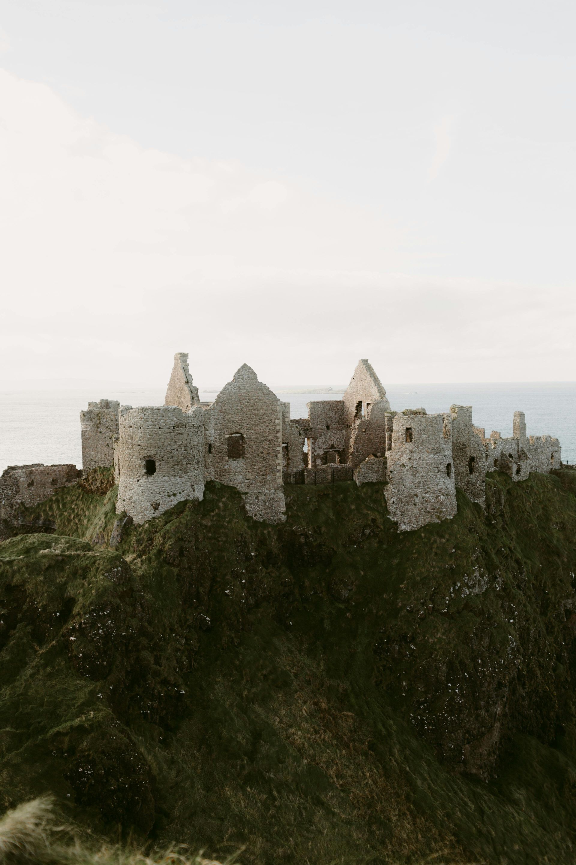 Ruined stone castle atop a grassy cliff overlooking the sea under a pale sky.