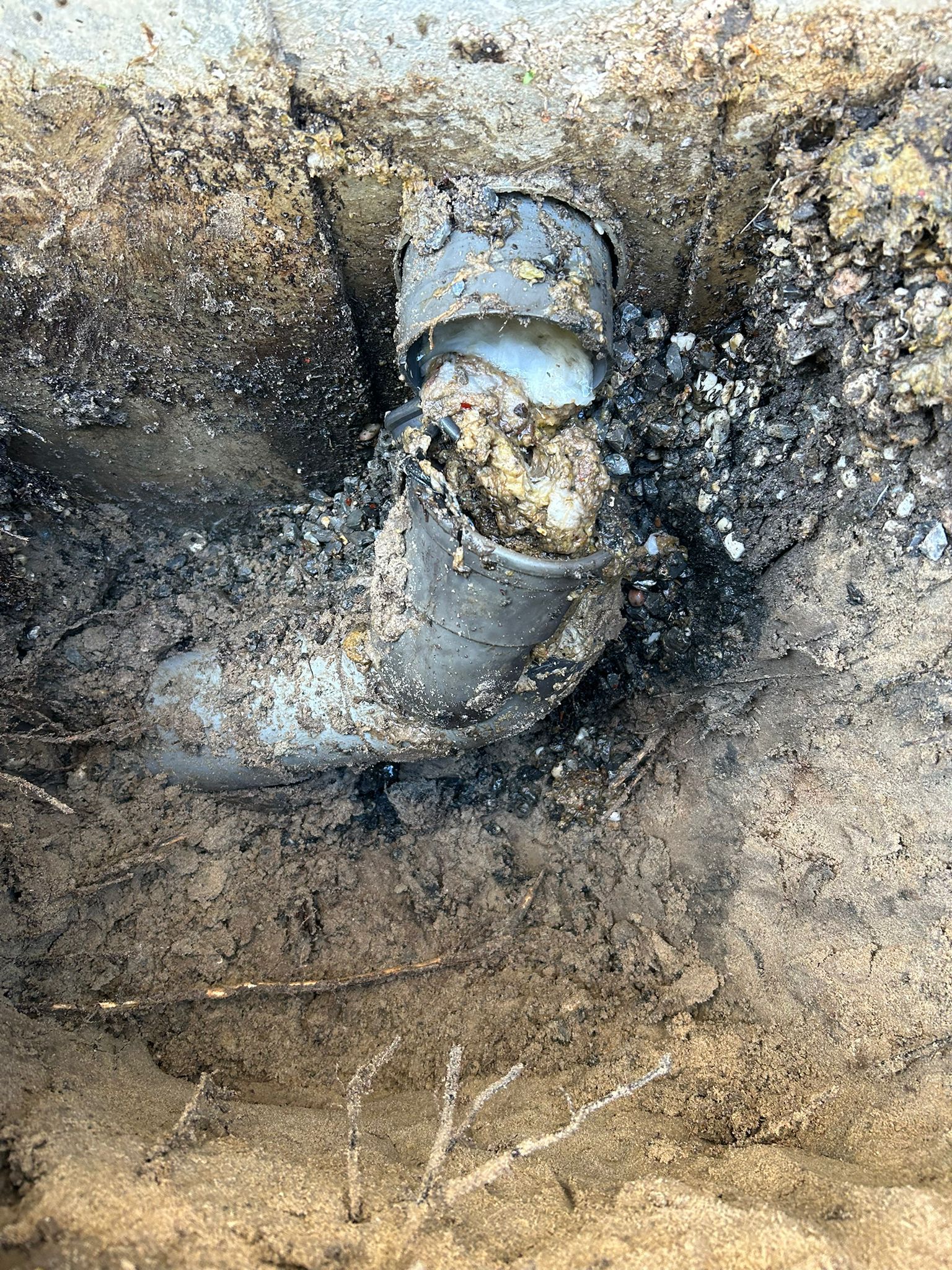 Broken pipe elbow exposed in muddy soil, with dirt and roots around it.