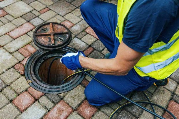 Utility worker cleaning a manhole on a brick pavement, wearing blue gloves and a yellow safety vest.
