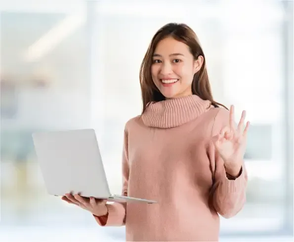 A woman in a pink sweater is holding a laptop computer and giving an ok sign.