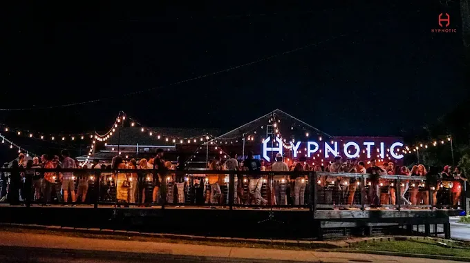 Hypnotic outdoor bar at night, with a crowd of people on a wooden deck, illuminated by string lights and a glowing sign.