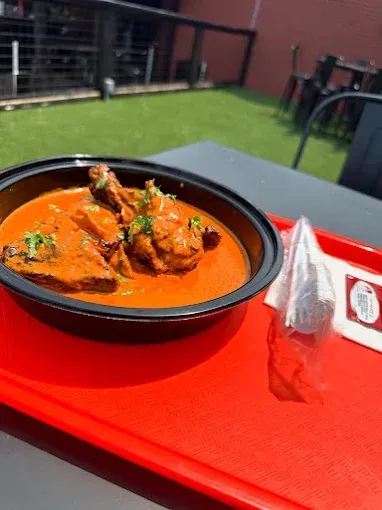 Chicken tikka masala with drumsticks in a black bowl on a red tray, outdoors on a sunny day.