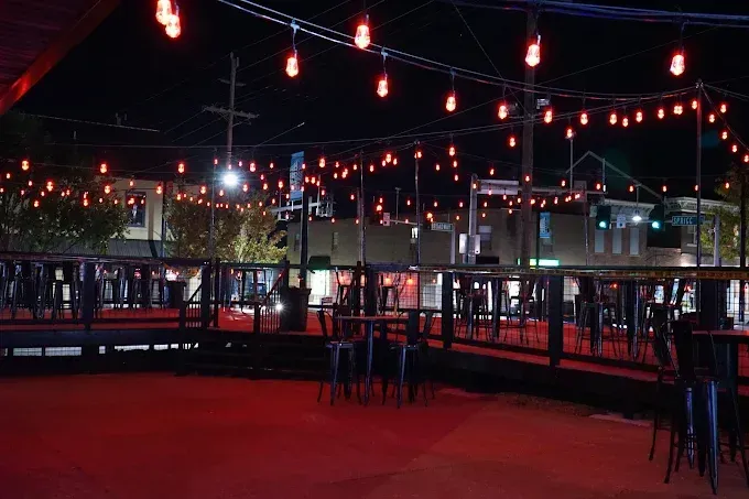 Nighttime outdoor patio with red string lights, tables, and stools. Buildings are visible in the background.