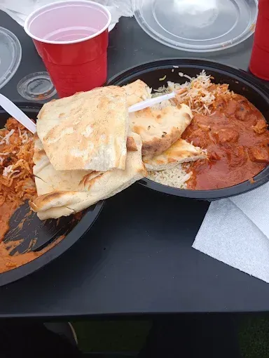 Two black bowls of Indian food on a black table, with naan bread, rice, and a red sauce. Red cups are on the table.