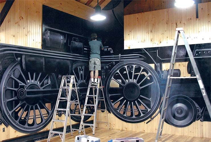 A man is standing on a ladder painting a train wheel on a wall.