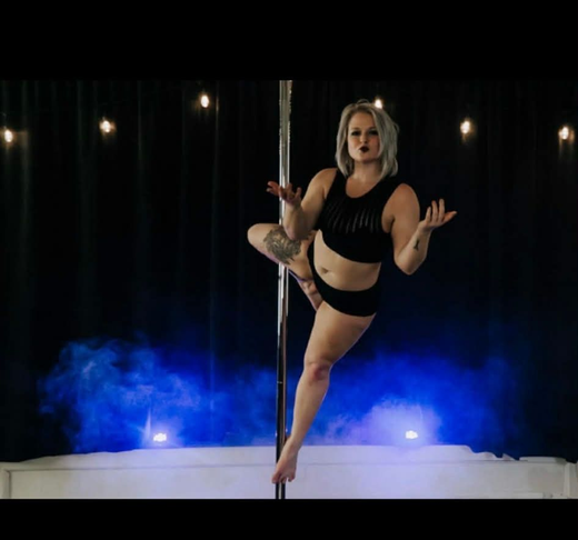 Pole dancer in a two-piece black outfit in a cupid pose with stage lights and smoke in the background