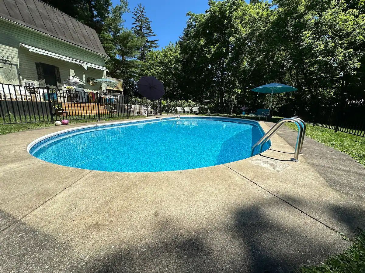An oval swimming pool with bright blue water sits in a backyard next to a light-colored house surrounded by trees.