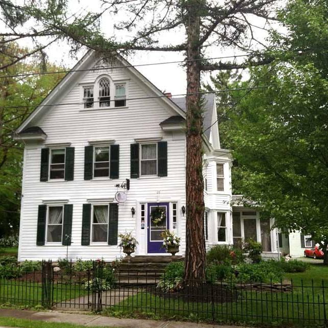 A two-story white house with dark green shutters, a blue front door, and a tall tree in the front yard.