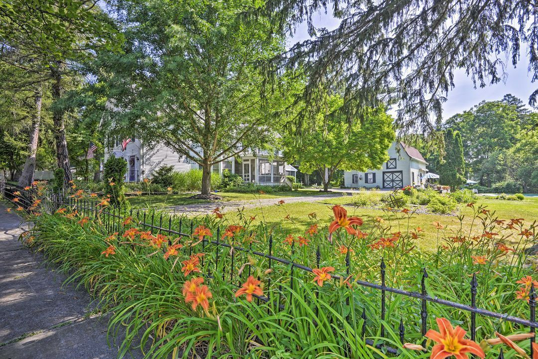 A row of orange daylilies behind a black metal fence in front of two white houses surrounded by mature green trees.