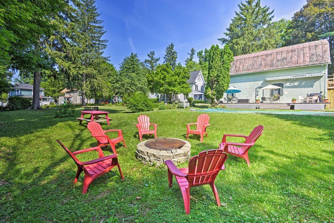 A stone fire pit surrounded by red Adirondack chairs in a grassy backyard near a light-colored house.