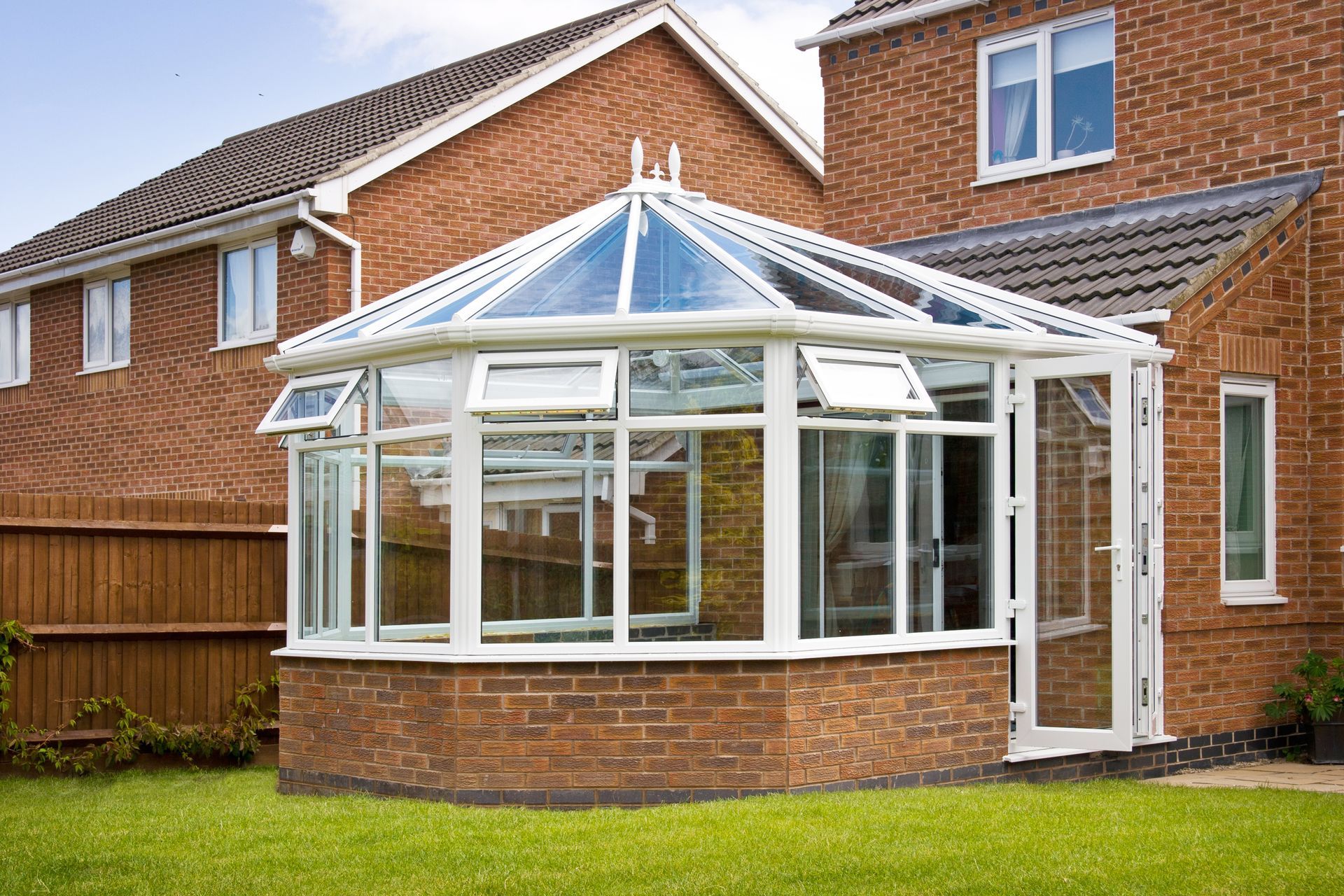A glass-roofed conservatory with white frames attached to a brick house, on a green lawn.