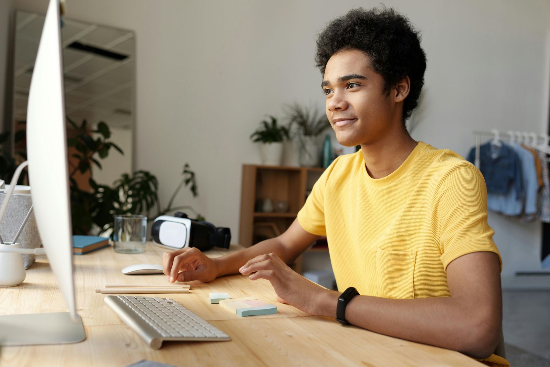 Person smiling at computer in a bright room, wearing a yellow shirt and black watch, wooden desk.