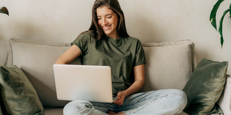 Young woman sitting on couch, smiling while using a laptop. She wears a green shirt and jeans.