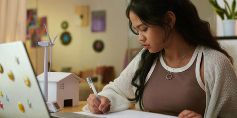 Girl writing at desk with laptop, small model wind turbine and house in focus.