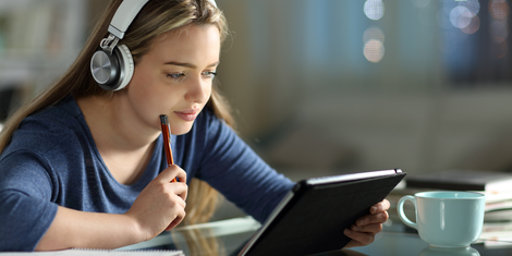 A young woman with headphones, using a tablet and pen while studying. Indoors.