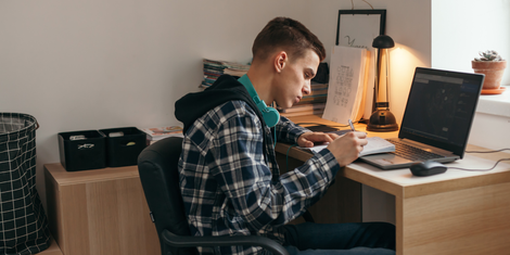 A young man studies at a desk with a laptop and notebook. He wears a plaid shirt, and headphones are around his neck.