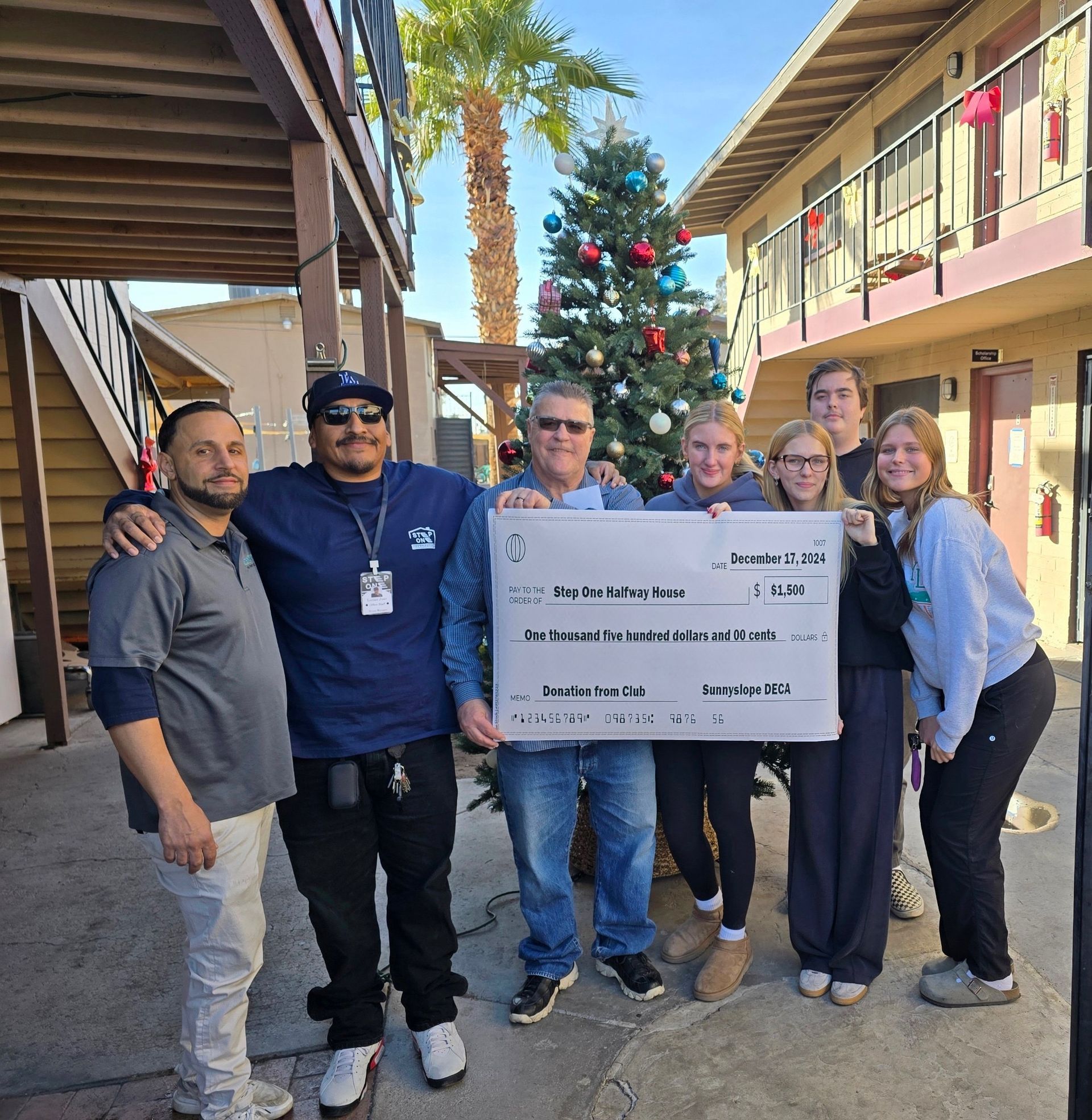 Group of people holding large check in front of Christmas tree and apartment buildings.
