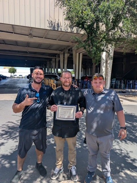 Three men outside, under a structure; one holds a certificate, others smile and point.