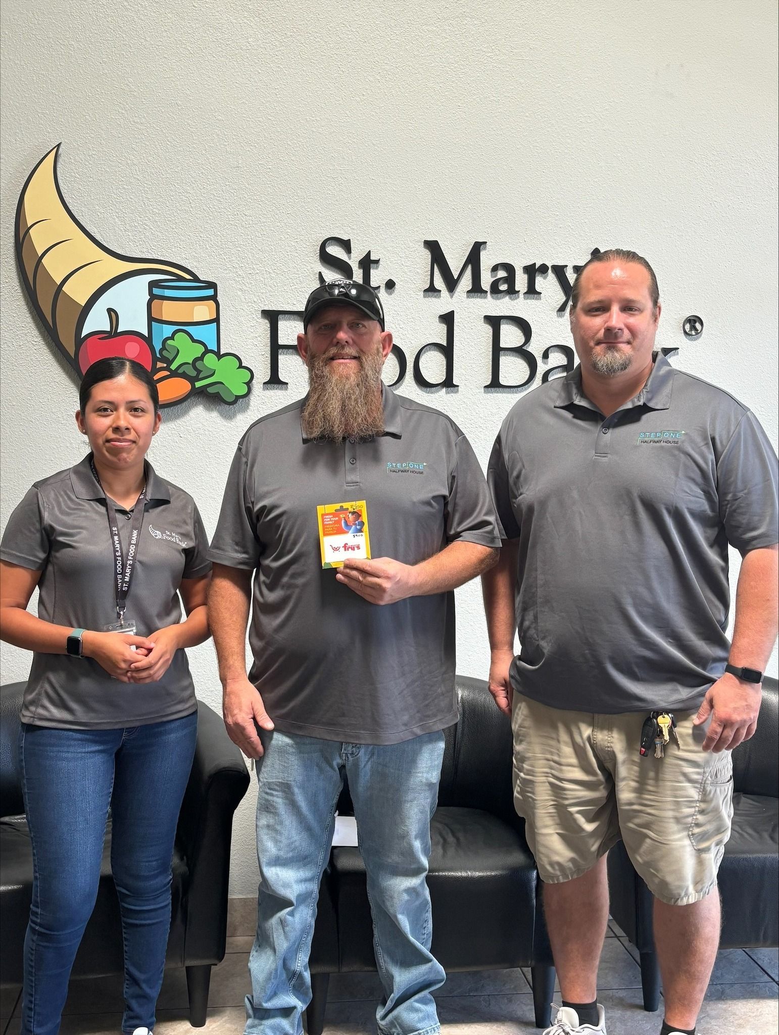 Three people at St. Mary's Food Bank; man holding a product, wearing gray shirts and standing in front of a logo.