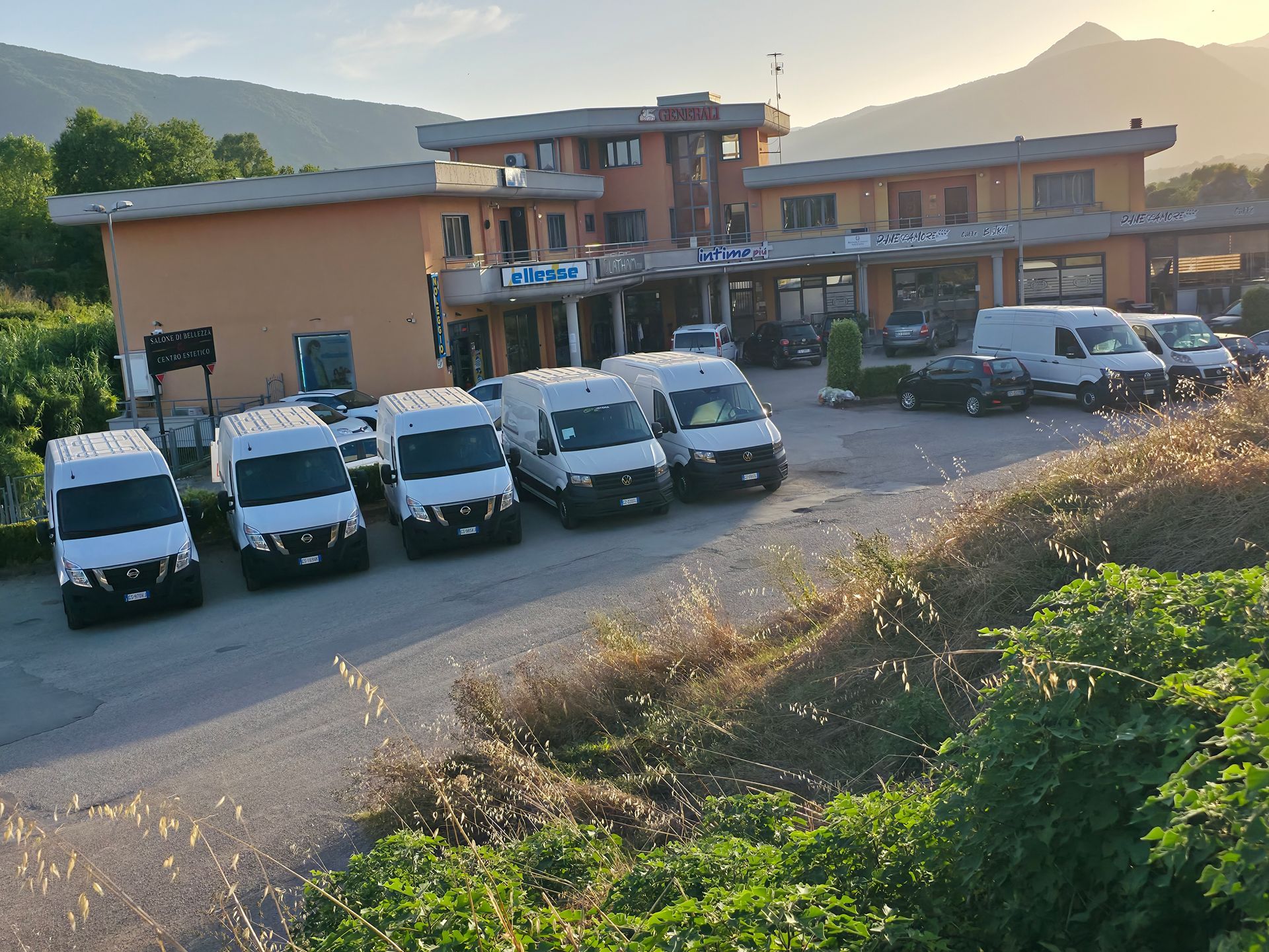 Una fila di furgoni bianchi parcheggiati davanti a un edificio color pesca in un contesto rurale di montagna al tramonto