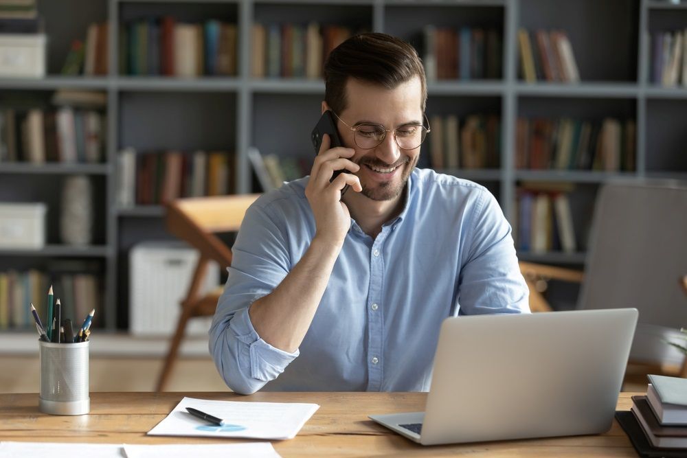 A Man is Sitting at a Desk With a Laptop and Talking on a Cell Phone — Repo Busters Wyong In Wyong, NSW