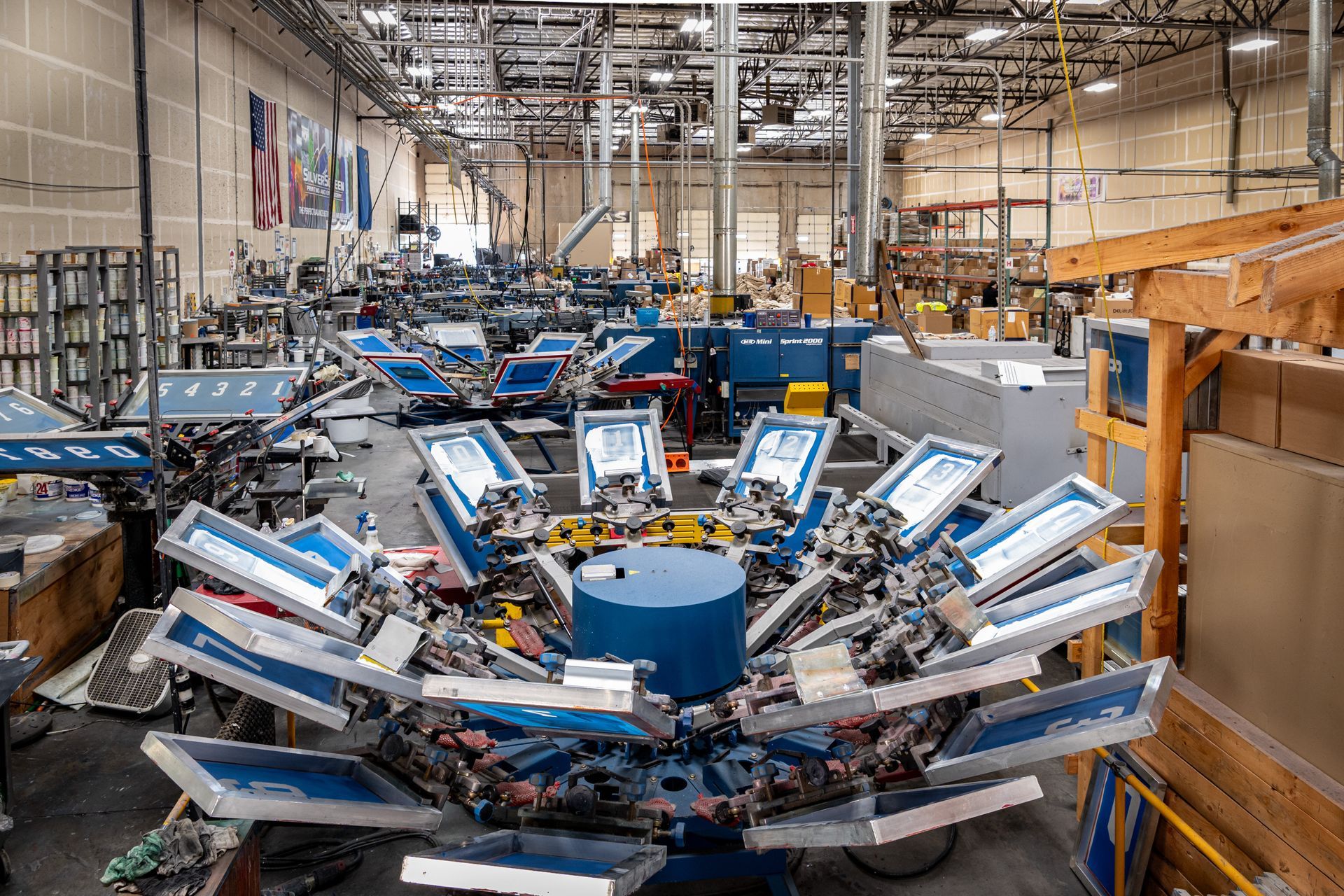 Screen printing machines in a factory. Many machines arranged in a workshop, under bright fluorescent lights.