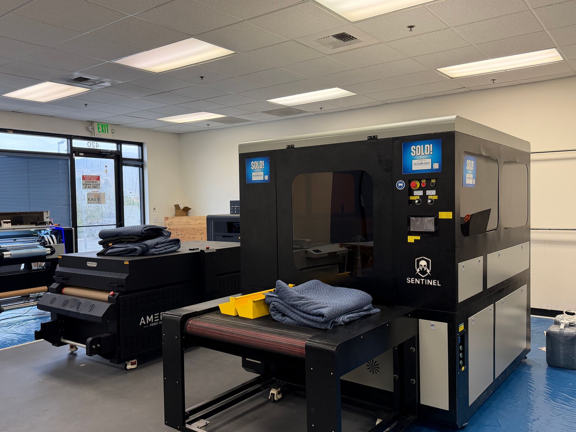 A large industrial DTF printing machine stands in a bright room with blue flooring, next to a conveyor belt with fabric.