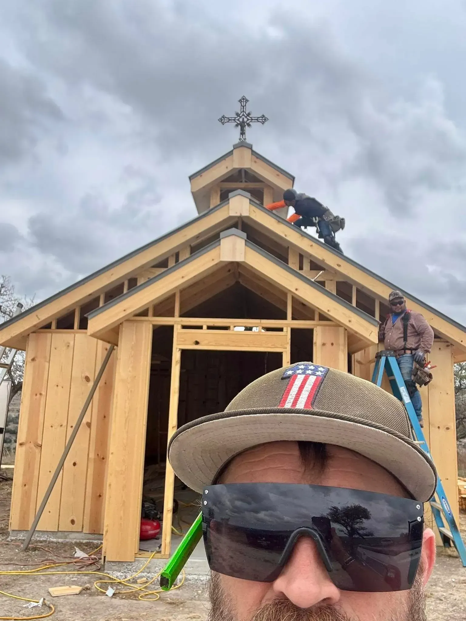 A man in a hat takes a selfie in front of a small chapel under construction, two workers on the roof.