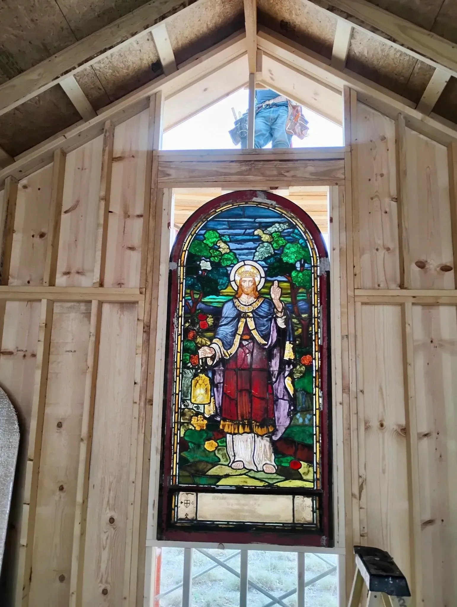 Inside a wood-framed structure, a stained glass window depicting a figure in a garden is installed. A worker is visible outside.