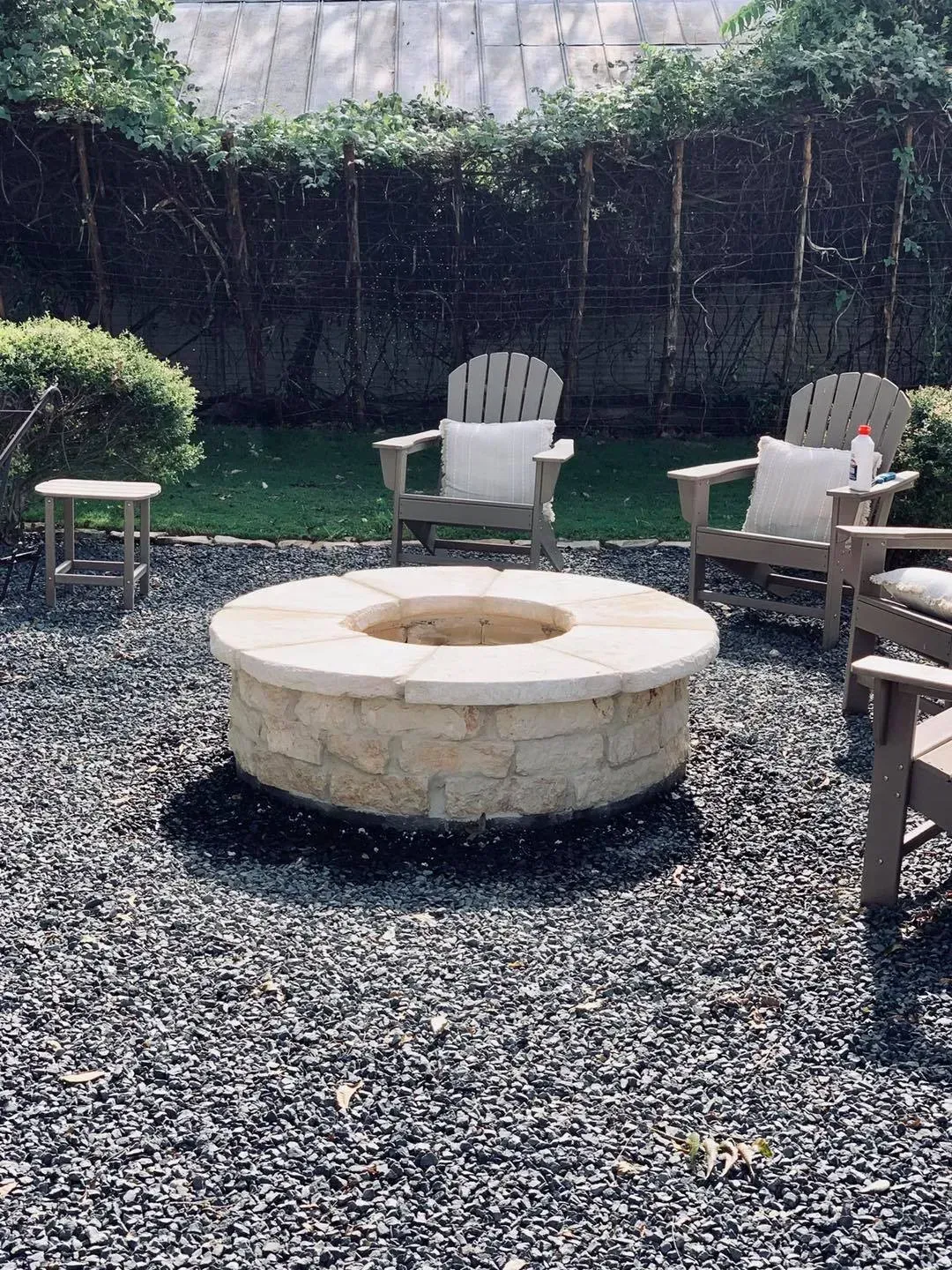 Fire pit in a gravel patio with Adirondack chairs, green grass, and overgrown background.