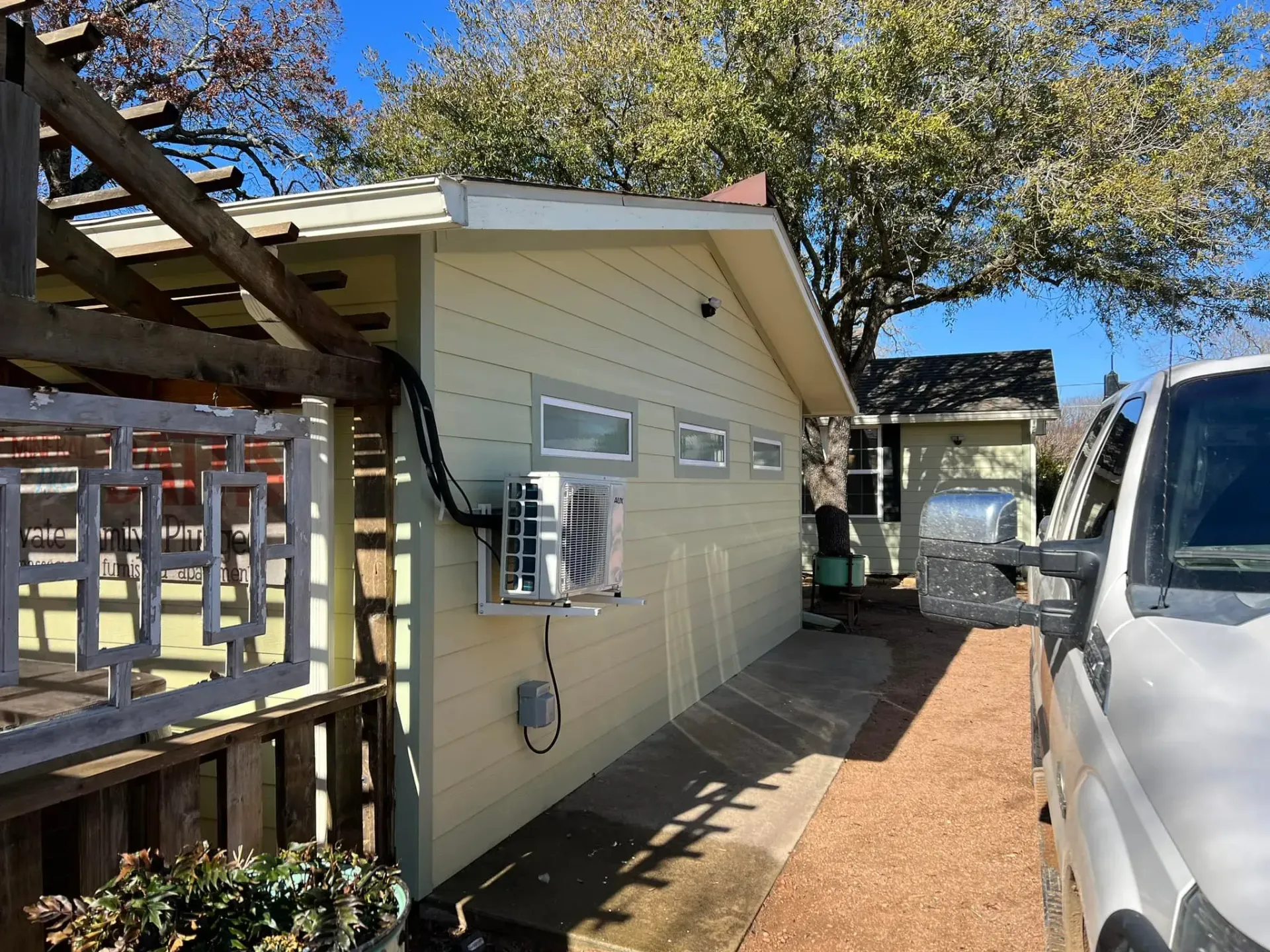 Tan-sided building with mounted AC unit, walkway, and parked white van. Pergola on left, trees in background.