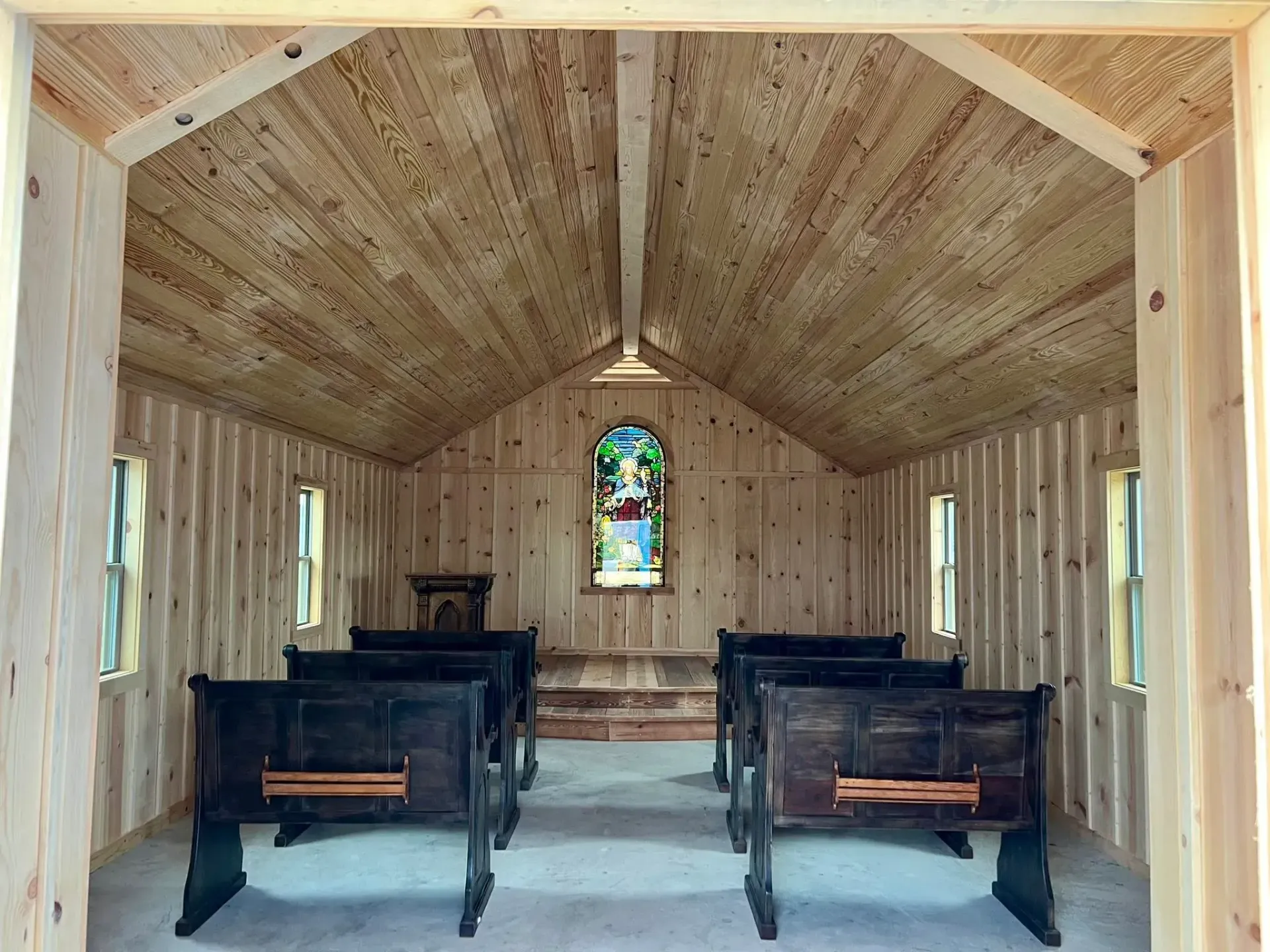 Interior of a small chapel with wooden walls, ceiling, and pews. A stained-glass window is centered at the altar.