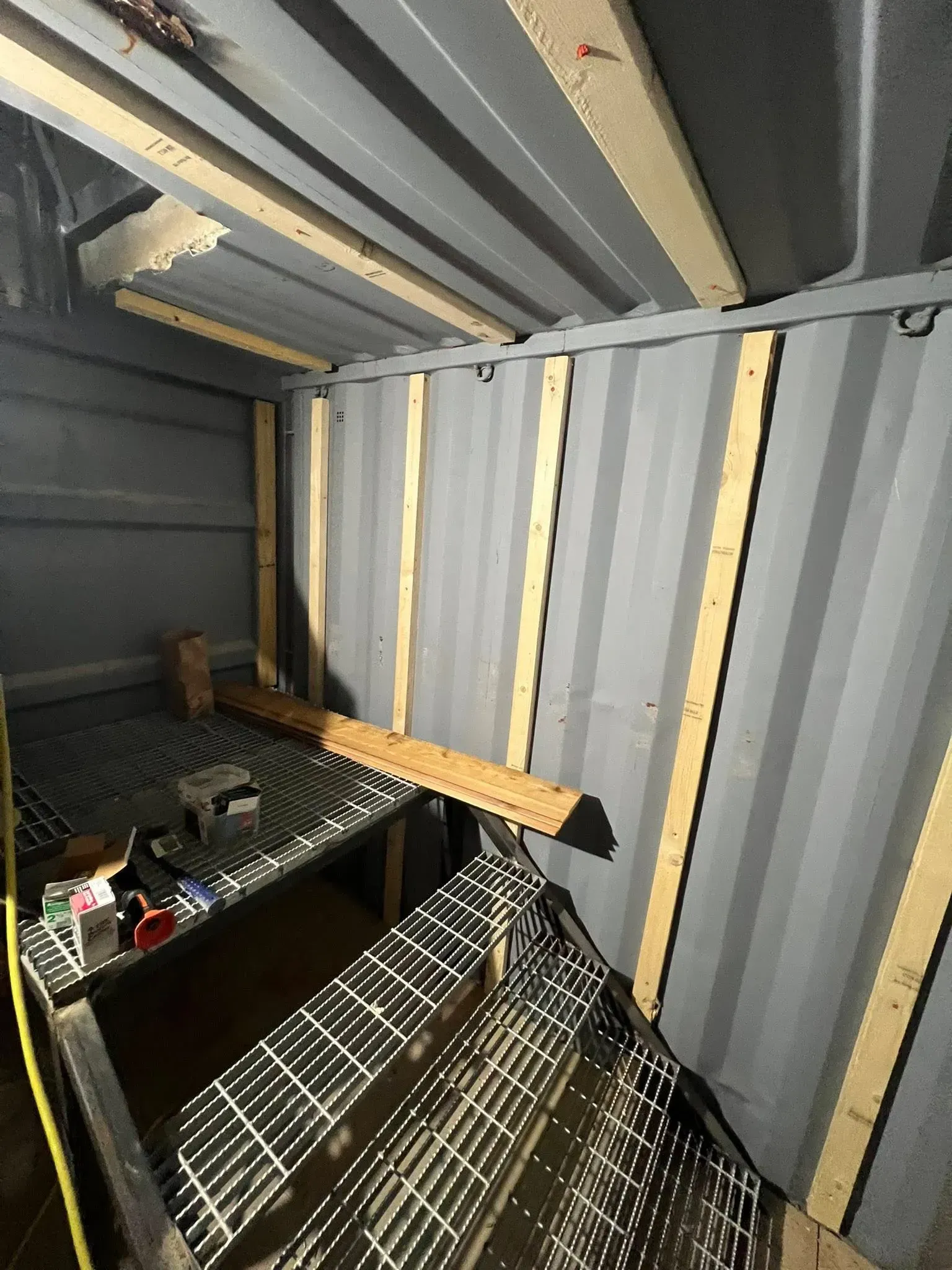 Interior view of a shipping container with metal grating, wooden supports, and overhead beams.