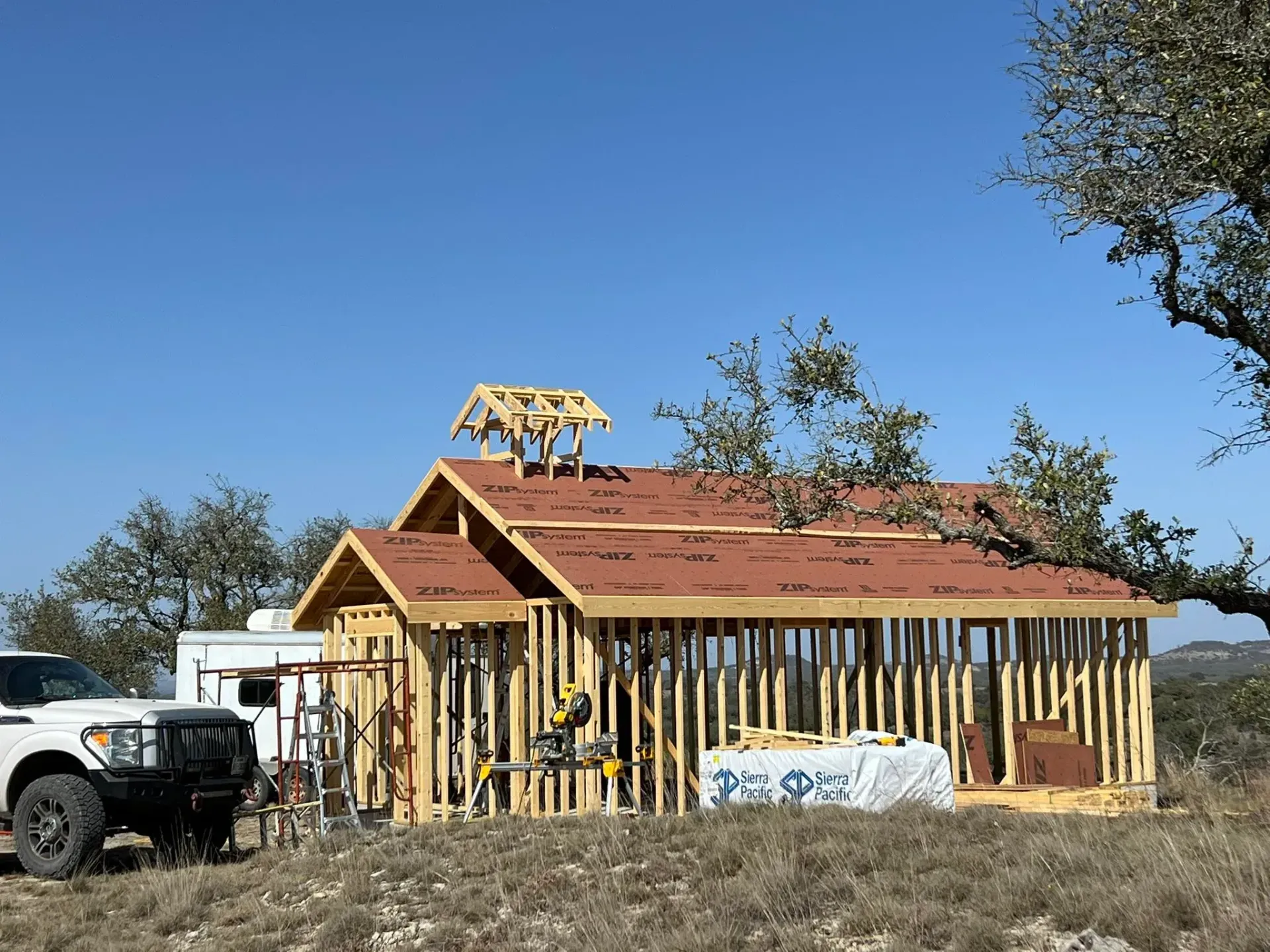Construction of a wooden shed with an unpainted roof, tools, and a white truck parked nearby, against a blue sky.