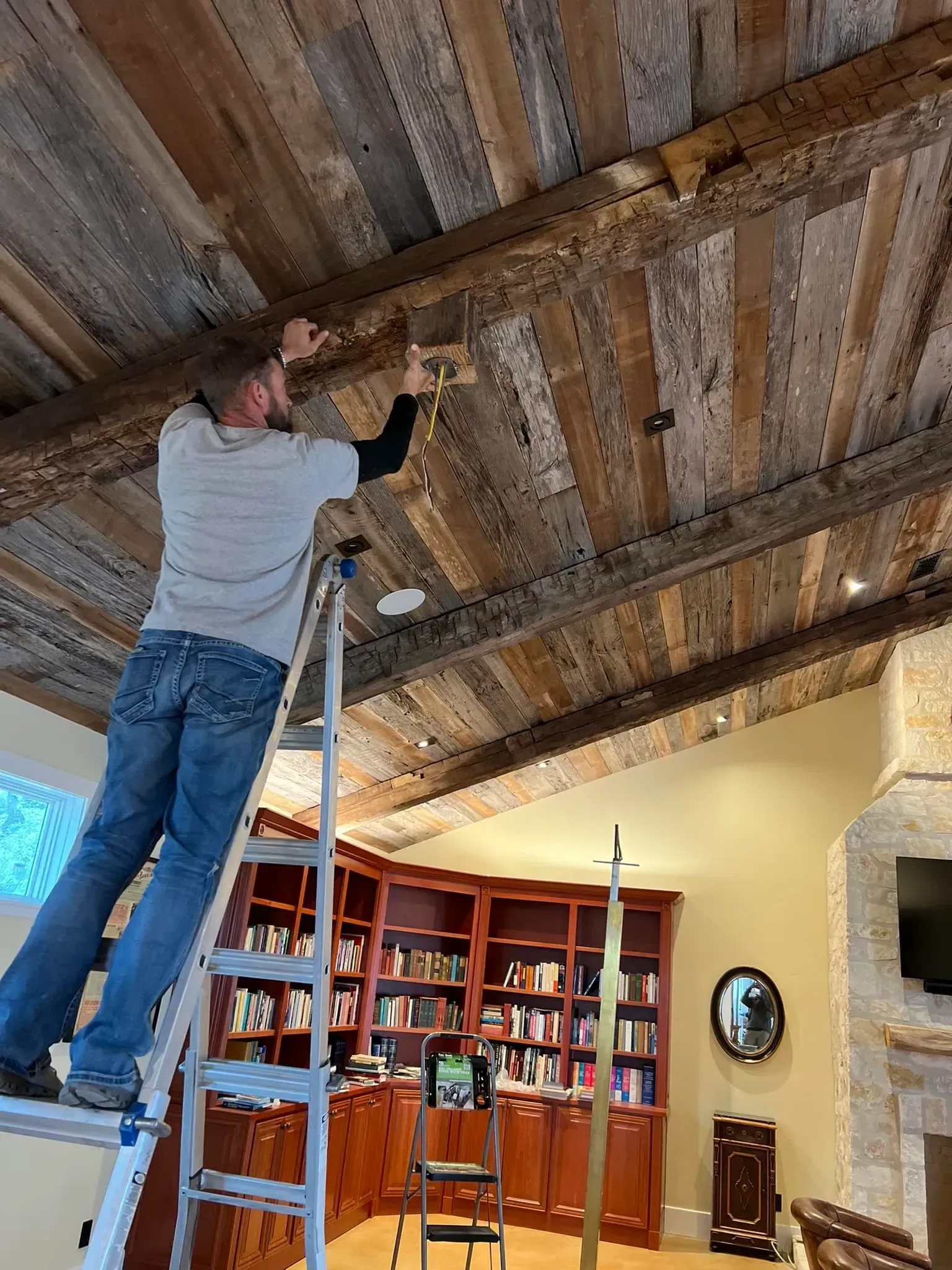 Man on ladder installing light fixture on wood plank ceiling.