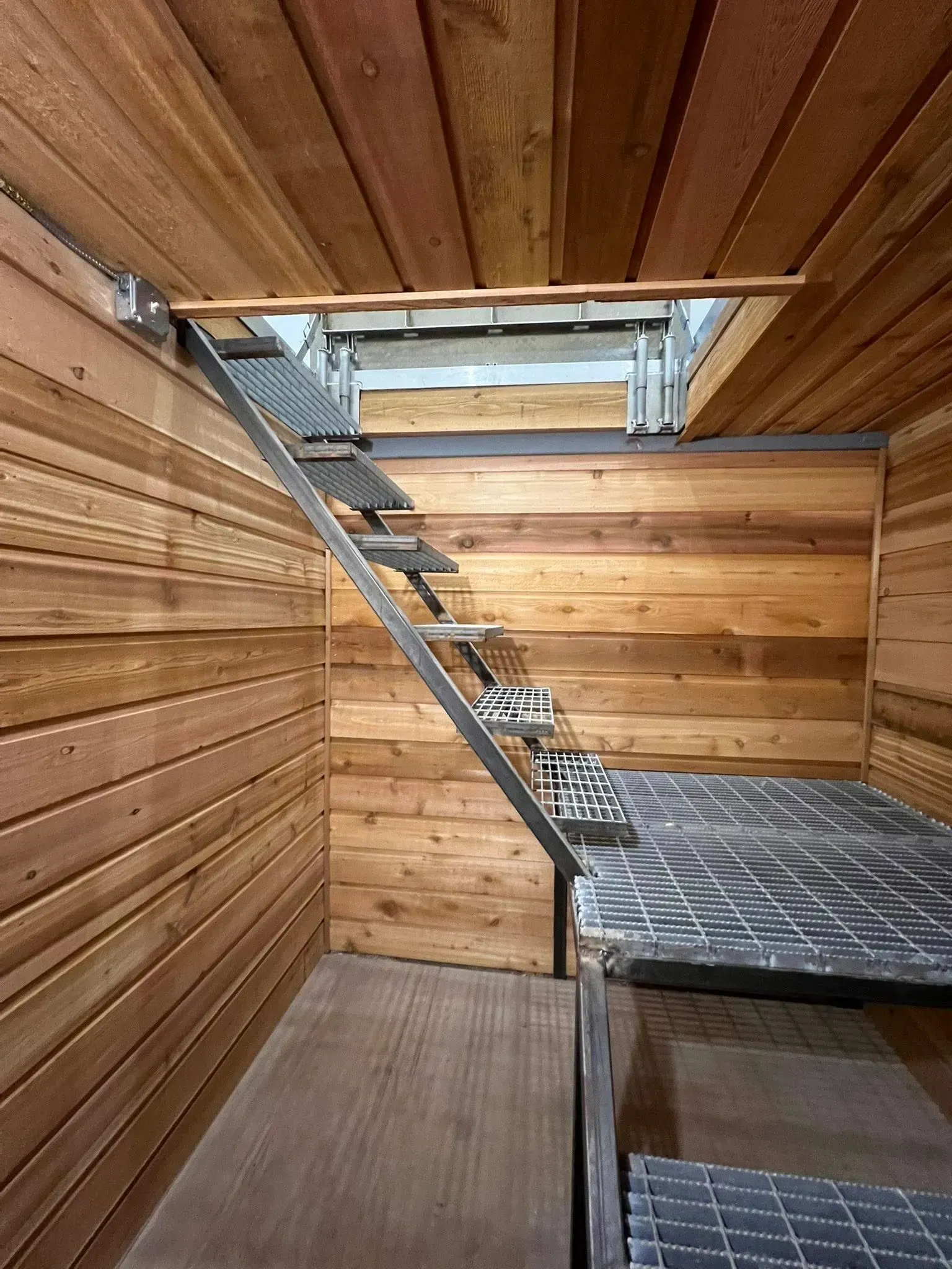 Interior view of a small room with wooden walls and ceiling. Metal ladder leads to an opening in the ceiling.