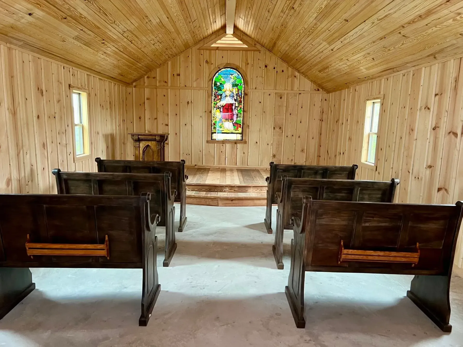 Interior of a small chapel. Wood-paneled walls and ceiling, pews, and a stained glass window.