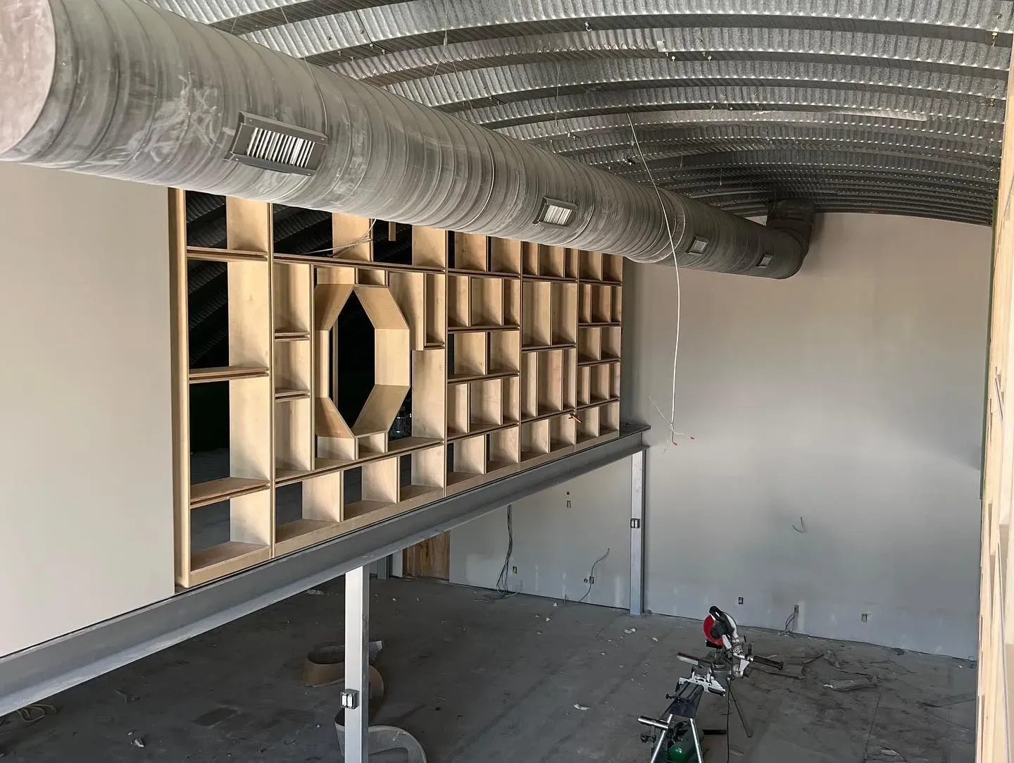 Interior view of a building under construction, featuring a wooden wall with shelves and a large air duct.