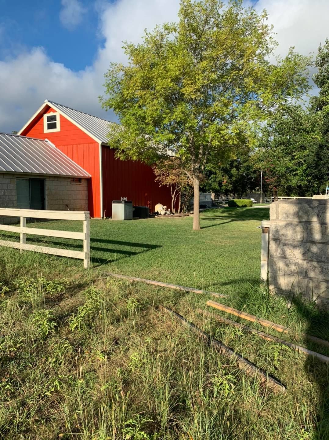 Red barn with metal roof and white fence in a grassy yard under a tree on a sunny day.