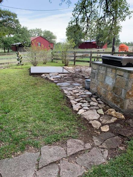 Stone pathway leading to a patio and grill in a grassy backyard, red barn in the distance.