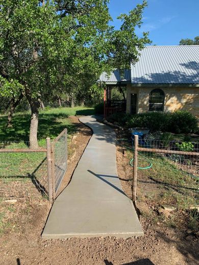 Newly poured concrete walkway leading to a house, with a small fence and gate. Sunny day.
