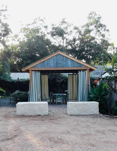 Outdoor gazebo with corrugated metal roof, curtains, and two stone benches. Green trees in background.