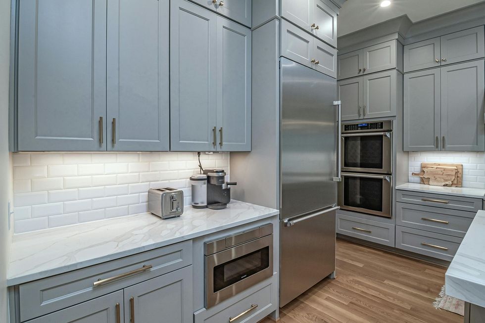A modern kitchen with grey cabinets, white marble countertops, a subway tile backsplash, and stainless steel appliances.