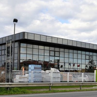 Edificio moderno con grandi vetrate che riflettono il cielo, dietro una recinzione bianca; materiali da costruzione visibili all'esterno.