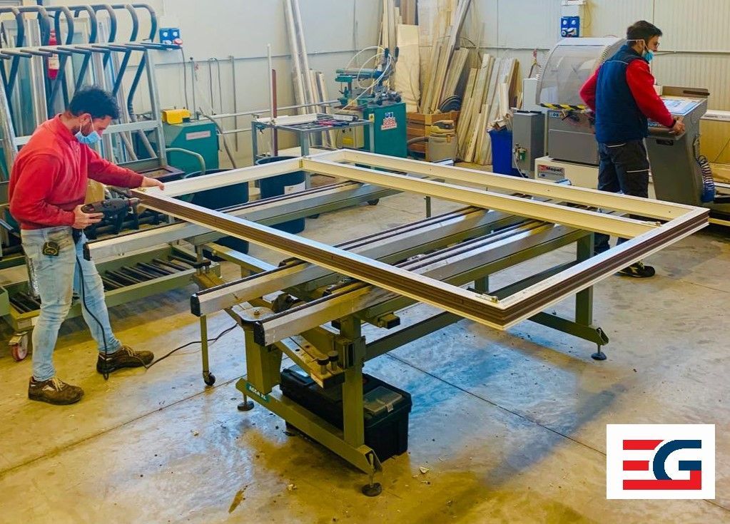 Due operai assemblano il telaio di una finestra in un'officina. Uno misura, l'altro aziona i macchinari.