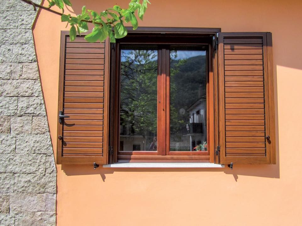 Finestra con persiane marroni aperta su una parete arancione, sulla quale si riflettono alberi e un edificio.